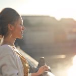 A young woman stands on a bridge enjoying the warm sunset over the city skyline.