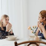 Mother and daughter share a cozy meal indoors, bonding and smiling at a dining table.
